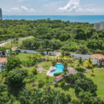Aerial view of Casa Dos Hermanos in San Carlos, with a curved blue pool in a green lawn among tropical trees and the ocean on the horizon.