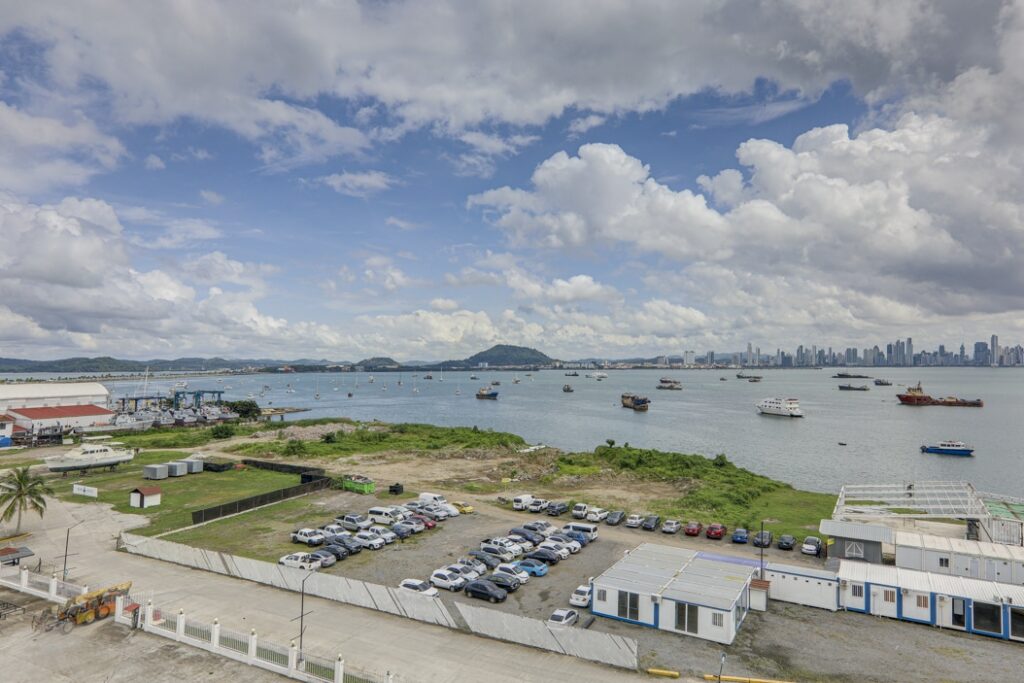 Harbor view over a crowded parking lot and boats on the bay, with a dense skyline across the water under a cloudy blue sky.