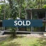 Modern two-story glass house surrounded by tropical foliage, overlaid with a SOLD banner.