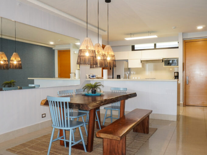 Dining area at Perlamar Casamar with a live-edge table, blue spindle chairs, woven pendant lights, and a white kitchen beyond.