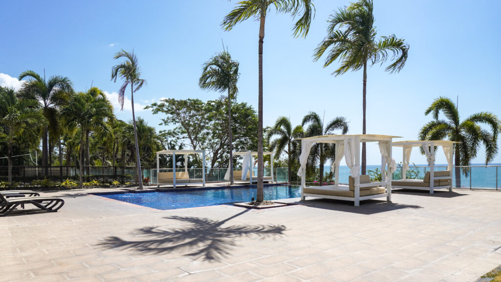 The pool deck at Palmar Residence in San Carlos has a blue tiled pool, white cabanas, and palm trees overlooking the ocean through a glass railing.