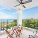 Image of balcony with two chairs, a table and chairs and views of the green spaces and ocean in this condo in Galeones