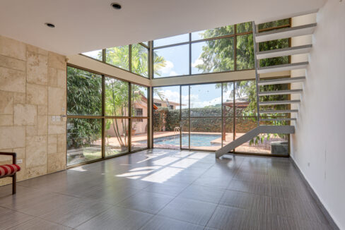 Empty room at La Alameda in Betania with gray tile floors, a white staircase, and floor-to-ceiling windows looking onto a patio pool.